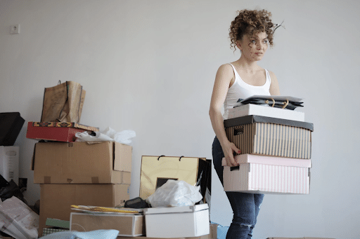 A woman carrying striped shoe boxes away from a pile of packed belongings in suitcases and shopping bags.