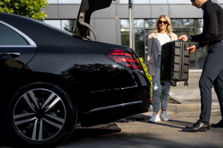 Businesswoman waits while chauffeur packs a suitcase in car trunk