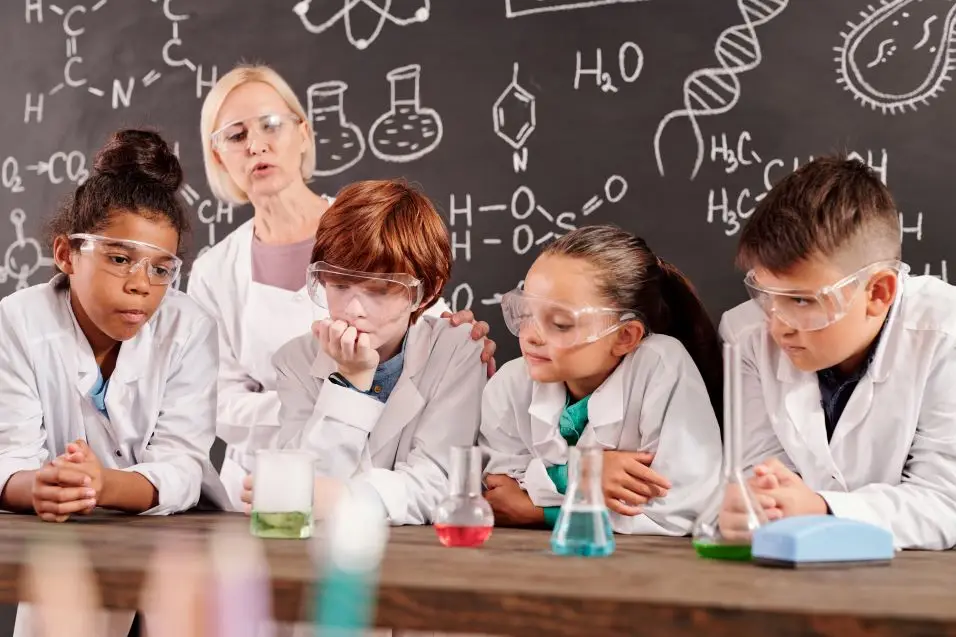 Group of glass tubes with chemical compounds standing in row on teacher desk in chemical laboratory while schoolkids watching different reactions